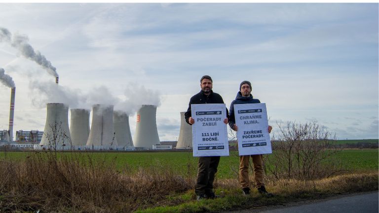 Dva lidé drží protestní cedule a v pozadí kouří komíny Počerad.